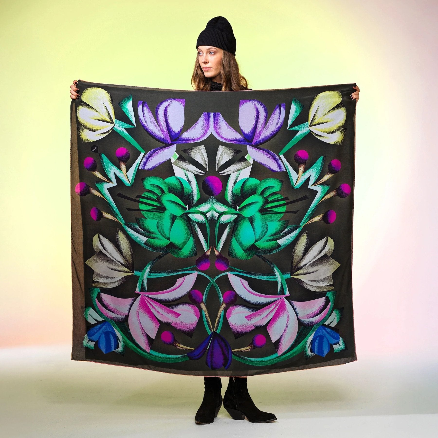 Frau mit schwarzer Mütze präsentiert grosses Tuch mit buntem, floralen Muster auf dunklem Hintergrund Damen Schweizer Design