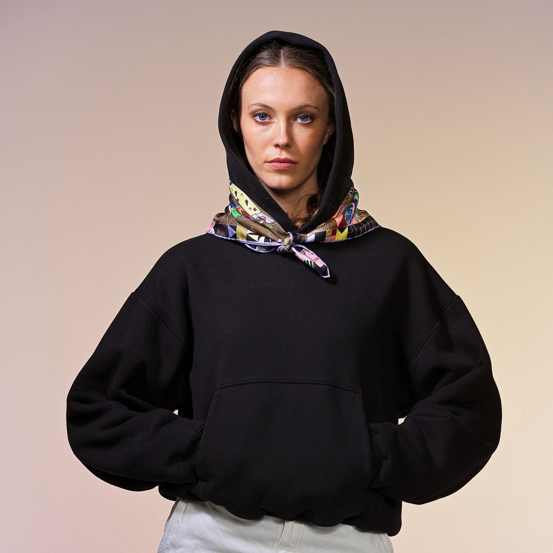 Frau mit schwarzer Kapuzenpullover trägt ein khaki Nicki-Seidentuch mit buntem abstraktem Muster, Stadtmotiv, CITY-AMA Design, quadratisch.