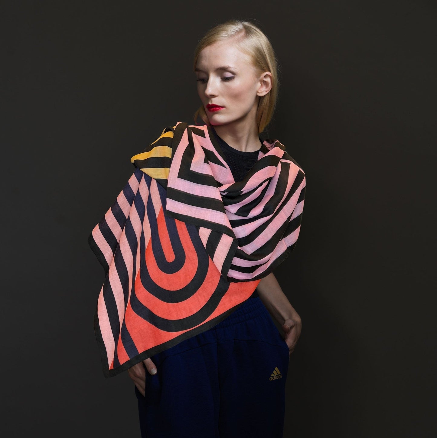 Frau mit blauen Jogginghosen trägt quadratisches Wolltuch mit gelben, rosa und roten Flächen und schwarzen grafischen Linienmustern