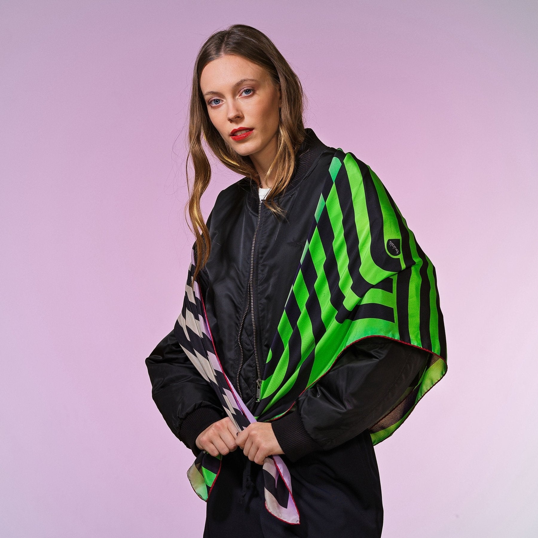 Frau mit schwarzer Bomberjacke trägt quadratisches Tuch mit neon-grünen und lila grafischen Linienmustern, Hintergrund lila-pastell