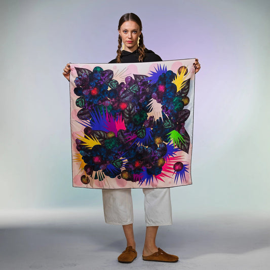 Frau hält buntes Foulala SAUVAGERIE Mimosa Tuch mit abstrakten Blumenmuster vor neutralem Hintergrund. Ein Seidencarré mit wildem Blumenmuster in Rot, Lila, Blau und Grün.