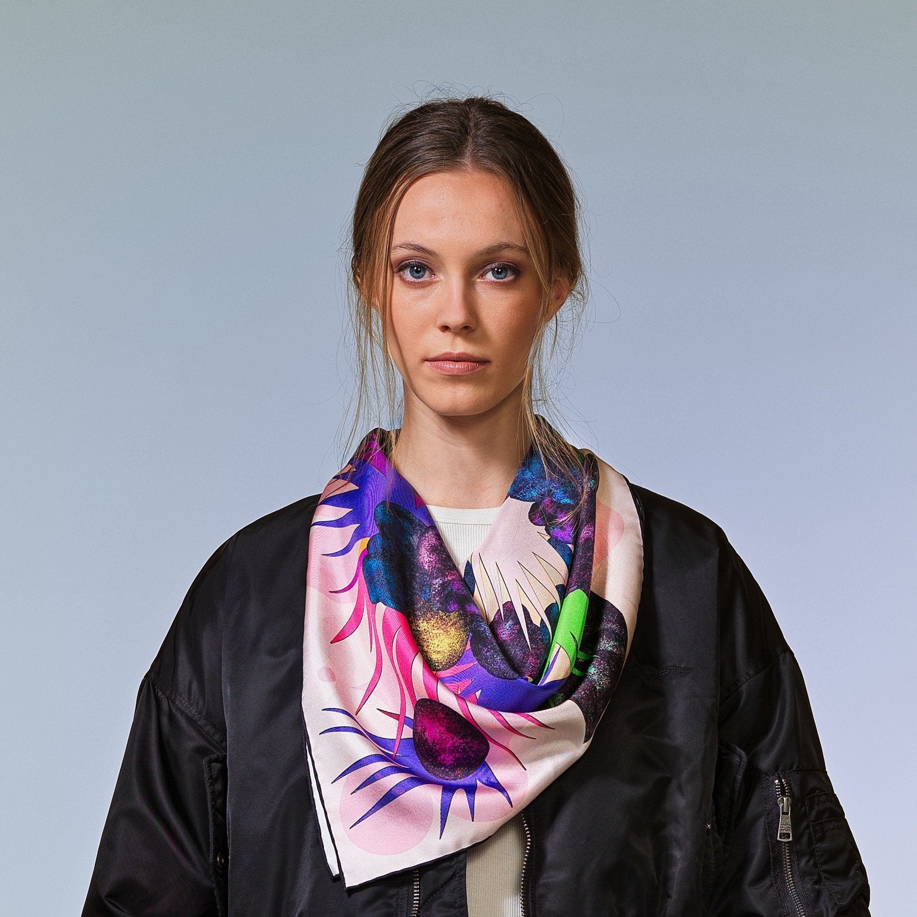 Frau mit brauner Bomberjacke trägt 
bunt gemustertes Seidentuch vor neutralem Hintergrund