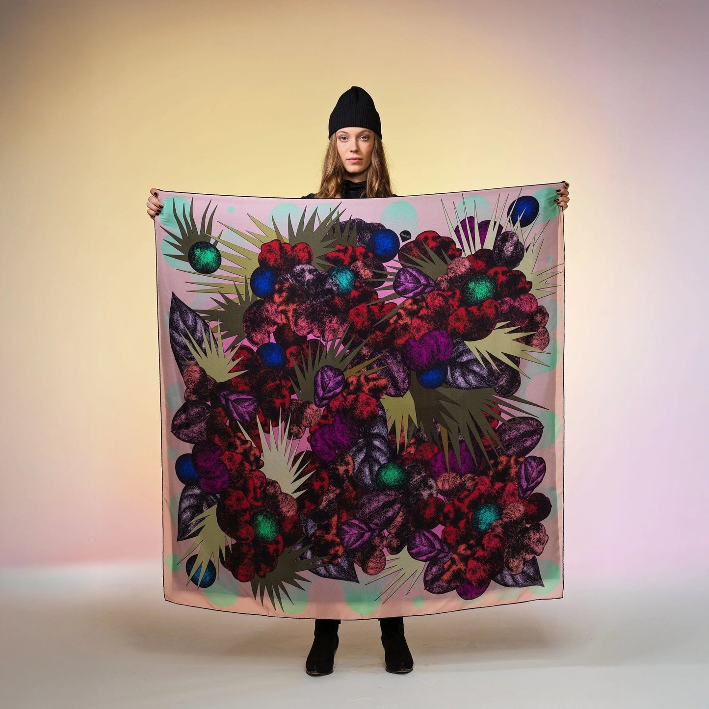 Frau mit schwarzer Mütze hält grosses, bunt gemustertes Seidentuch Foulard von Foulala vor neutralem Hintergrund